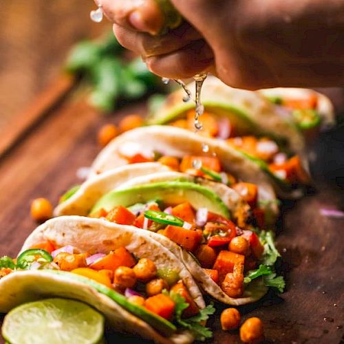 The image shows a hand squeezing lime over a row of tacos filled with chickpeas, vegetables, and garnished with cilantro on a wooden surface.