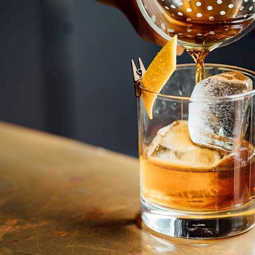 A hand pours a brown drink into a glass with ice and a lemon twist garnish on a countertop.