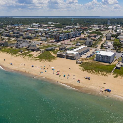 Aerial view of a beach with clear water, scattered umbrellas and beachgoers. Residential buildings and greenery are visible in the background.