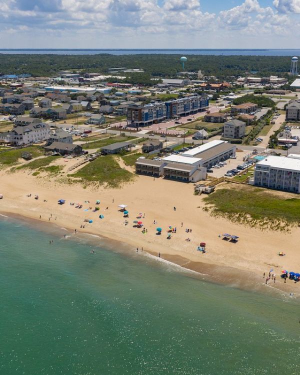 Aerial view of a beach with clear water, scattered umbrellas and beachgoers. Residential buildings and greenery are visible in the background.