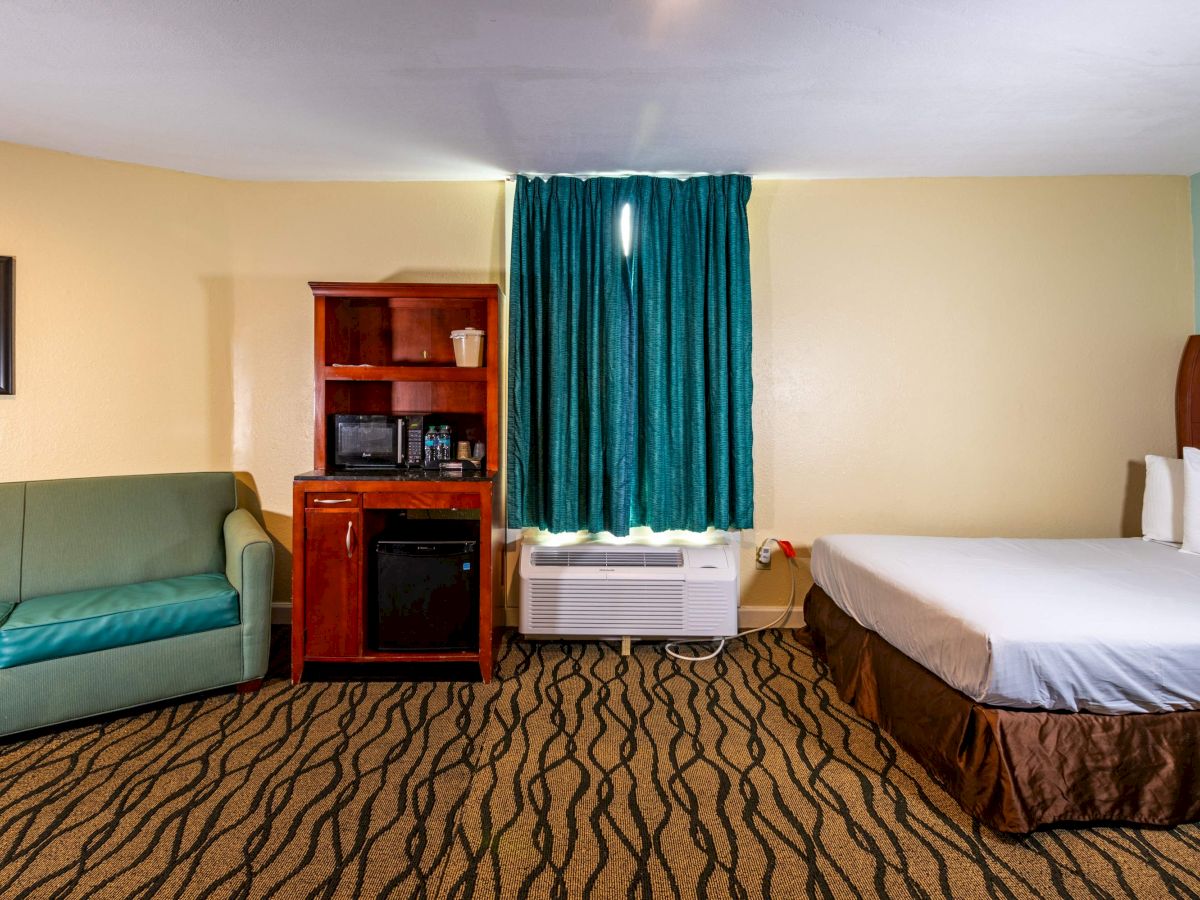 The image shows a hotel room with a bed, a green sofa, a cabinet with a microwave and mini fridge, and a window with teal curtains.