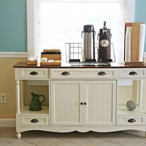 A white sideboard with coffee dispensers, cups, and napkins on top, set against a window with blinds in a teal and beige room.