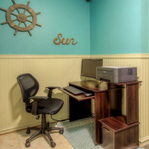 A small office area with a computer, printer, and chair. Decor includes a helm and the word 