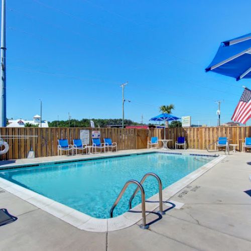 A sunny poolside scene with lounge chairs, umbrellas, and American flags, surrounded by a wooden fence and clear blue sky.