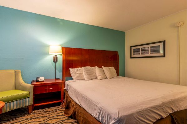 A hotel room with a bed, nightstand, lamp, framed pictures, and an armchair. The walls are blue and the furniture is wooden.