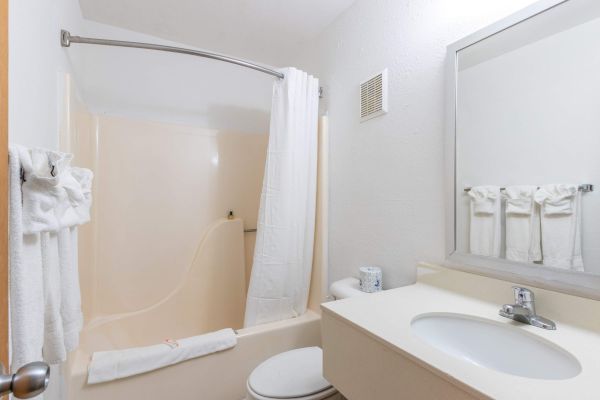 The image shows a small bathroom with a shower-tub combo, white curtain, sink, mirror, and towels. Bright lighting enhances the clean look.