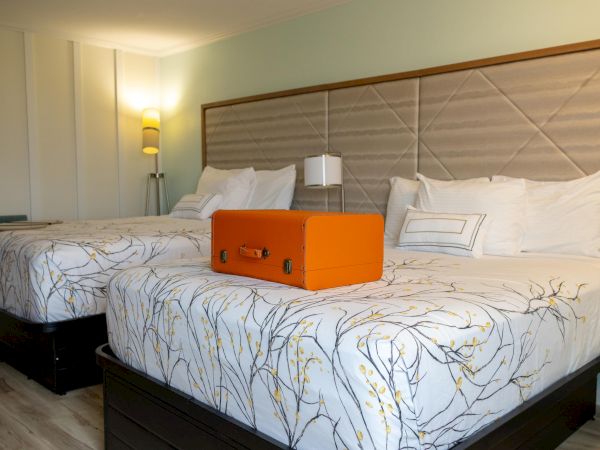 The image shows a hotel room with two beds, floral bedding, a bright orange suitcase on a bed, and modern lighting.