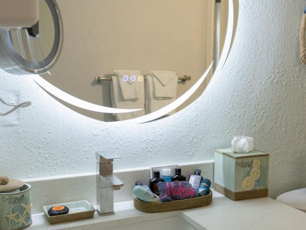 Modern bathroom with a round, backlit mirror. A sink with toiletries and tissues sits on the countertop beneath the mirror.