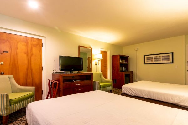 The image shows a hotel room with two beds, a TV on a dresser, chairs, a lamp, and artwork on the wall.