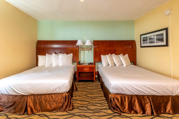 The image shows a hotel room with two double beds, a shared nightstand, lamps, pillows, and a wall picture with a patterned carpet.