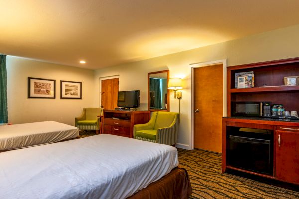 The image shows a hotel room with two beds, a TV, chairs, a small fridge, and wall art. The room is warmly lit and carpeted.