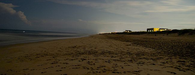 A serene beach scene at dusk with footprints in the sand and a few distant buildings illuminated by light in the background.