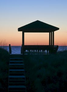 A silhouette of a gazebo overlooks the ocean at sunset, with steps leading up, against a colorful sky and dark foreground.