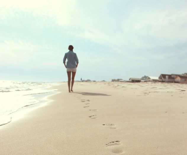 A person walks along a sandy beach, leaving footprints behind, with the ocean to their left and houses in the distance.