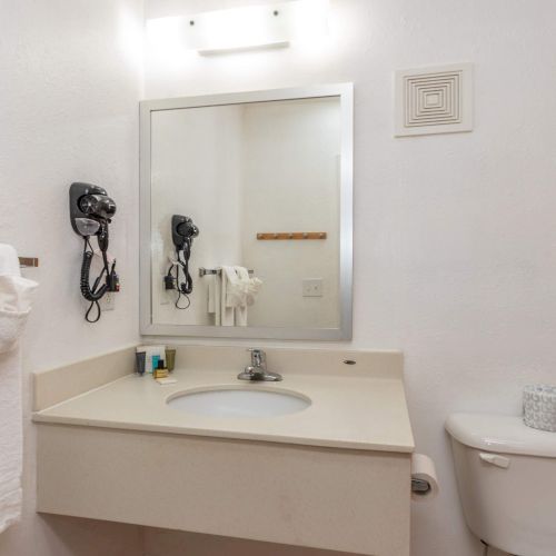 A bathroom with a vanity, mirror, wall-mounted hairdryer, towels, toilet, and a shower curtain, all in a white color scheme.