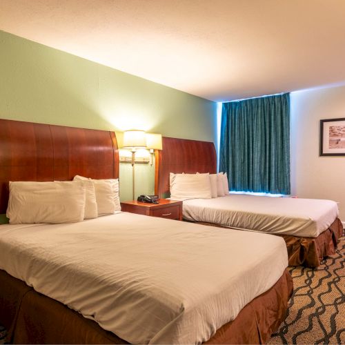 The image shows a hotel room with two double beds, a lamp, framed pictures on the wall, and patterned carpet, creating a cozy atmosphere.