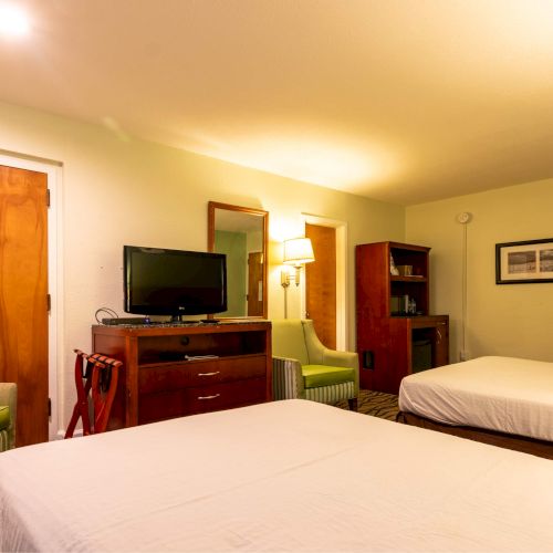 The image depicts a hotel room with two beds, a TV on a dresser, chairs, a small table, and framed artwork on the wall.