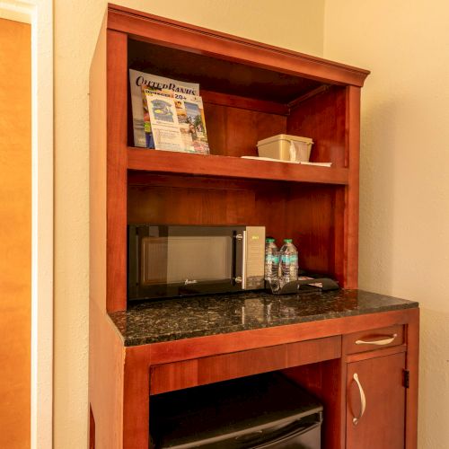 A wooden kitchenette with a microwave, water bottles, a brochure, and a mini-fridge below, topped with a black countertop.