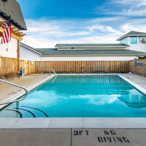 An outdoor swimming pool with clear water, surrounded by a wooden fence, a building, and a U.S. flag. The depth is marked as 3 feet.