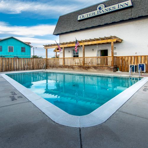 An outdoor swimming pool at Outer Banks Inn with a wooden fence and buildings in the background under a partly cloudy sky.