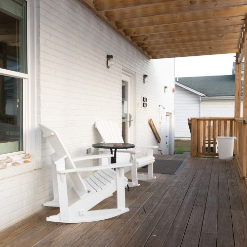 A porch with two white rocking chairs, a small table, and a wooden railing. A row of stones decorates the windowsill.