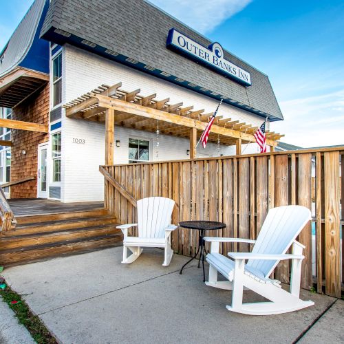 The image shows a building with a sign, a wooden ramp, two white chairs, and a small table outside, framed by a fence and clear sky above.