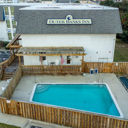 An aerial view of Outer Banks Inn featuring a fenced outdoor pool and adjacent buildings with parking area.