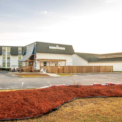 A building with a parking lot and landscaped area featuring mulch and small shrubs. The sky is partly cloudy in the background.