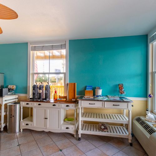 The image shows a bright room with turquoise walls, white cabinets, a coffee station, and large windows, offering a cozy, inviting atmosphere.