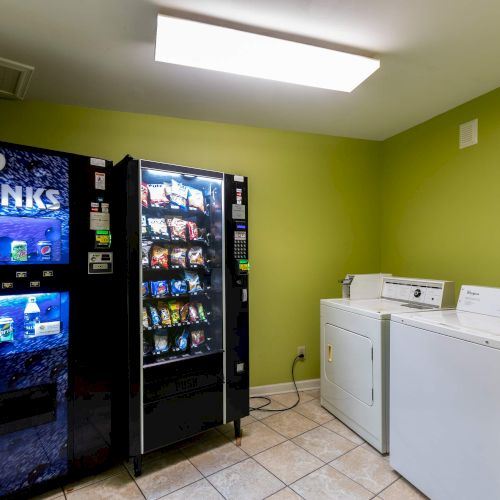 A room with a cold drinks vending machine, a snack vending machine, and a washer and dryer on a tiled floor with green walls.