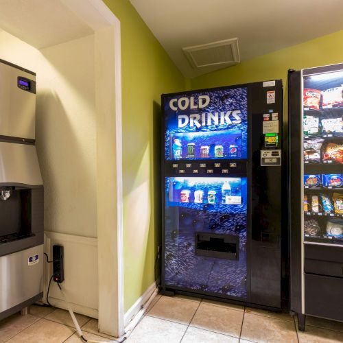 An ice machine, a cold drinks vending machine, and a snacks vending machine are placed side by side in a room with tiled flooring.