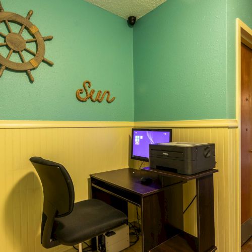 A small hallway corner with a desk, chair, computer, and printer. A ship wheel decor and 