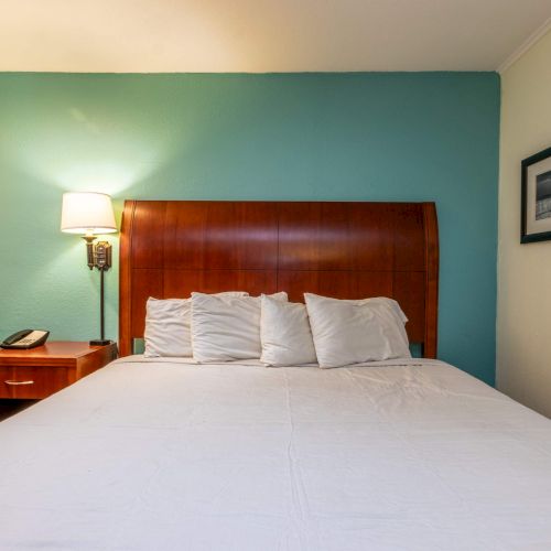 A hotel room with a bed and wooden headboard, a side table with a lamp and phone, a chair, and a framed picture on the wall.