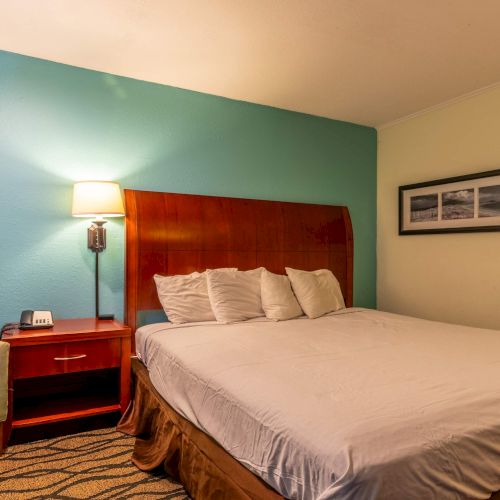 A hotel room with a large bed, green chair, side table, lamp, and framed artwork on a blue wall. The room has a simple, cozy design.