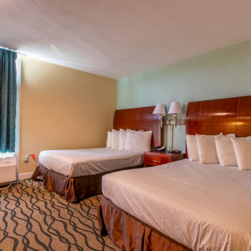 A hotel room with two beds, a side table with a lamp, and curtains covering a window. The carpet has a patterned design.