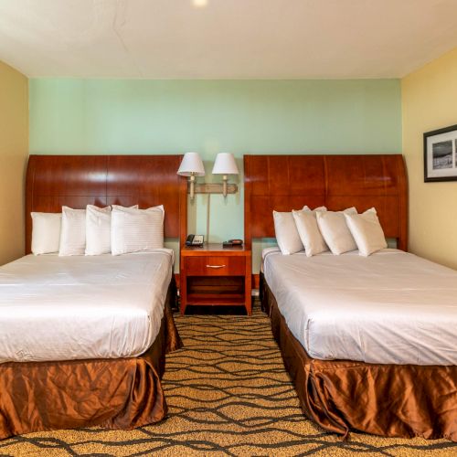 The image shows a hotel room with two double beds, four pillows each, a shared nightstand, lamps, and a framed picture on the wall.