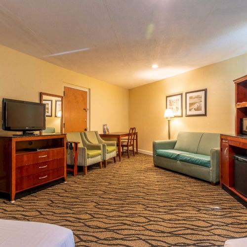 A hotel room with a bed, TV, green sofa, chairs, and a wooden cabinet. Carpeted floor and ceiling light enhance the cozy atmosphere.