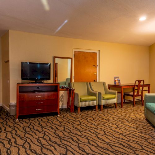 The image shows a hotel room with green chairs, a sofa, a TV, a table with chairs, a lamp, and a patterned carpet.