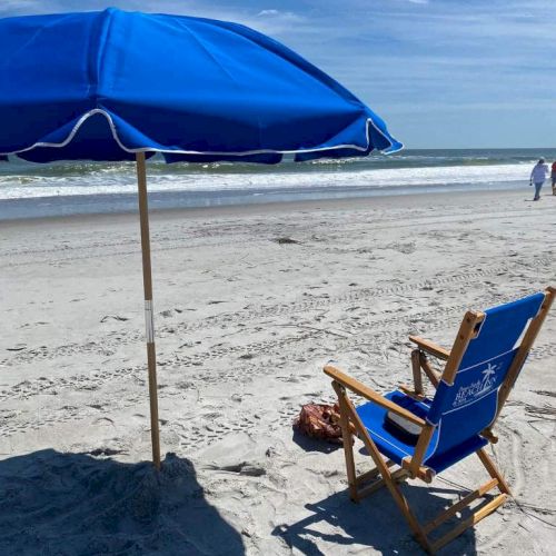 A beach scene with a blue umbrella and chair facing the ocean. A bag is nearby, and the sky is partly cloudy.