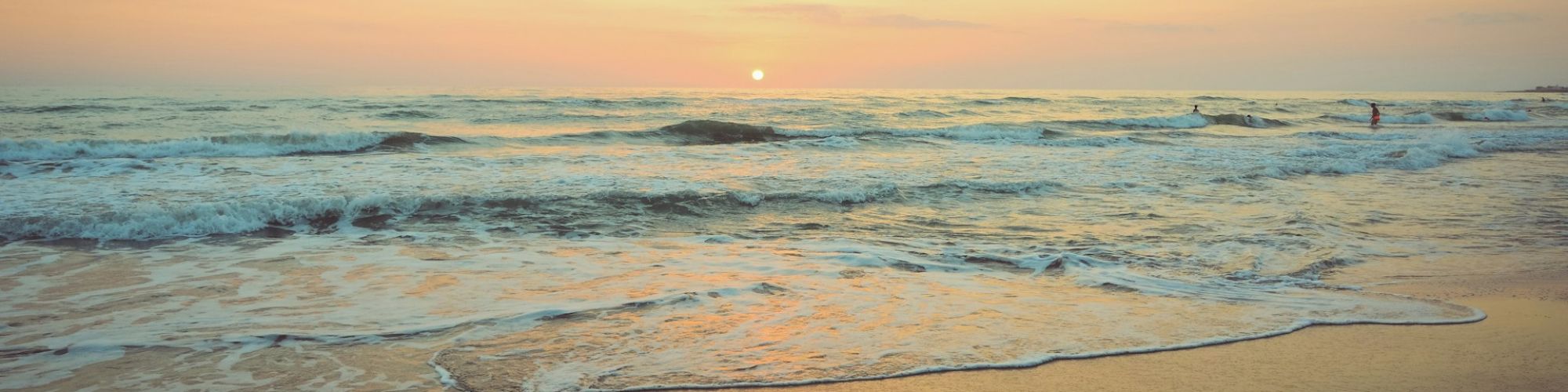 A serene beach scene at sunset with calm waves gently hitting the shore, under a sky painted in soft orange and pink hues.