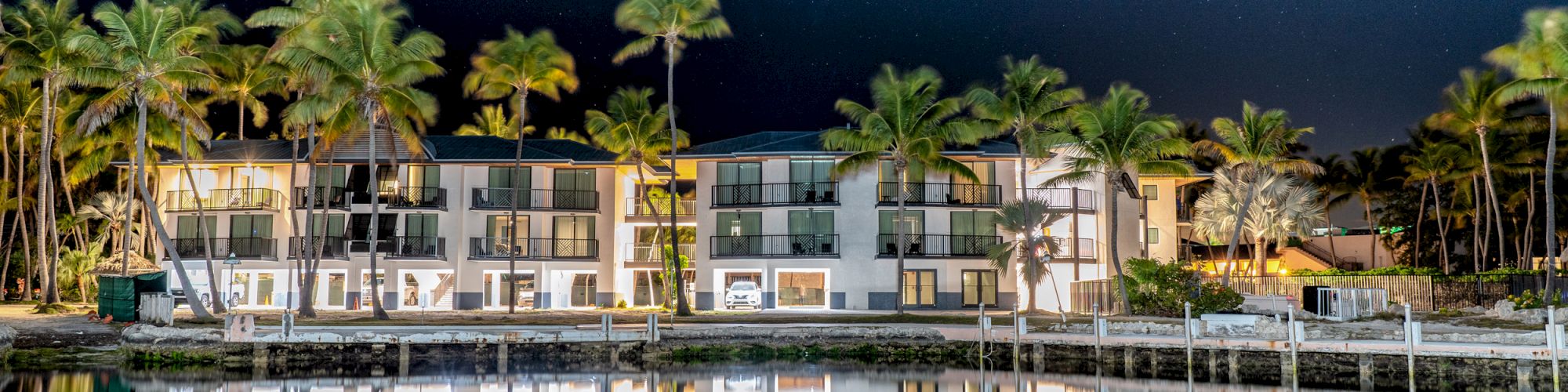 A luxurious waterfront hotel under a starry night sky, surrounded by palm trees, perfectly reflected in the calm water, creating a serene scene.