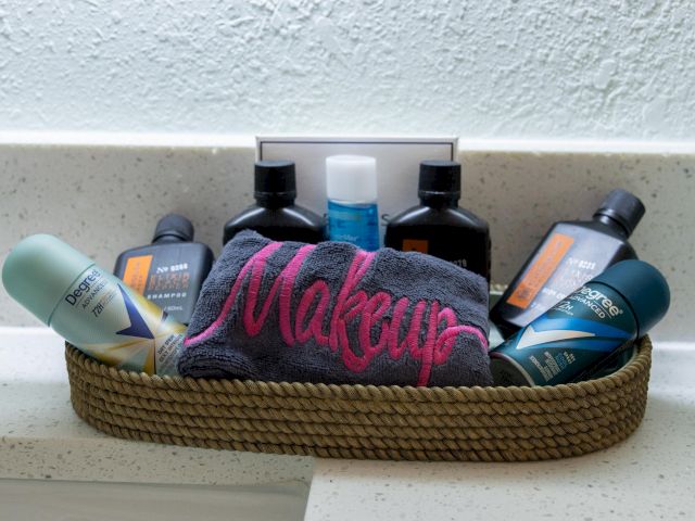 A basket contains toiletries, a small towel labeled "Makeup," various bottles, and deodorant, arranged neatly on a bathroom counter.