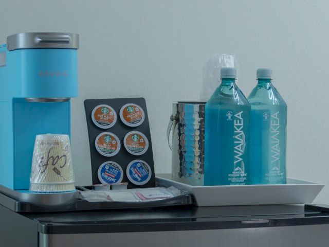 A coffee maker with pods, water bottles, cups, and a tray are placed on a countertop or mini-fridge.