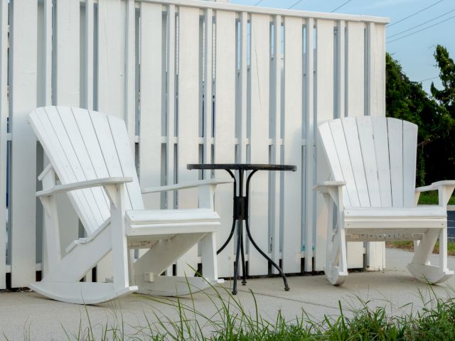 Two white Adirondack chairs with a small black metal table sit on a patio, backed by a white vertical plank fence.