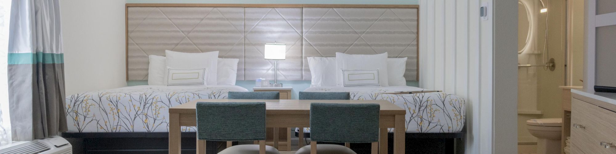 The image shows a modern hotel room with two beds, a table with chairs, and a view into a bathroom. The decor is minimal and clean.